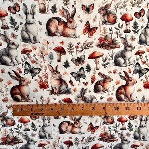 A close-up of fabric with a pattern of rabbits, mushrooms, butterflies, and autumn leaves, with a wooden ruler across the bottom.