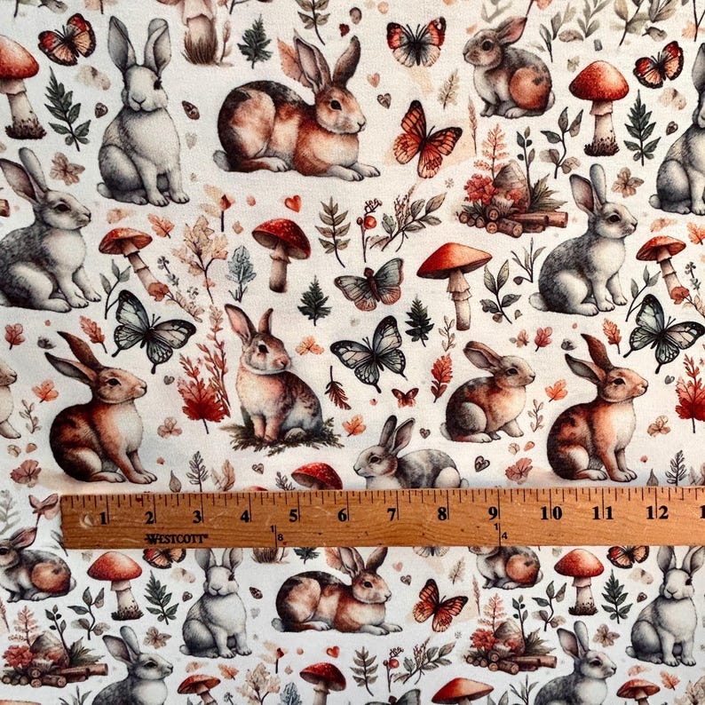 A close-up of fabric with a pattern of rabbits, mushrooms, butterflies, and autumn leaves, with a wooden ruler across the bottom.