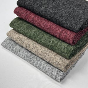 Op de afbeelding: Een stapel van vijf stofstalen in verschillende kleuren en patronen. De stalen zijn gemaakt van een zacht, getextureerd materiaal. De kleuren zijn grijs, beige, groen, bordeauxrood en zwart.