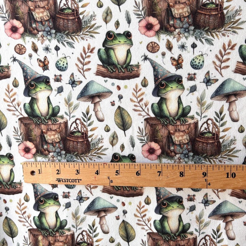 A ruler is laid across fabric displaying a repeating pattern of green frogs in wizard hats, mushrooms, and leaves on a white background.