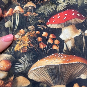 Close-up of dark woodland fabric featuring colorful mushrooms and ferns, with a hand with pink nail polish touching the left edge.