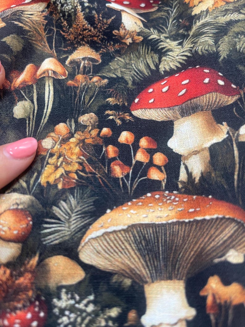 Close-up of dark woodland fabric featuring colorful mushrooms and ferns, with a hand with pink nail polish touching the left edge.