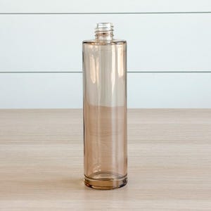 May include: A tall, cylindrical glass bottle with a light amber or champagne-colored tint. The bottle features a smooth, straight-sided body and a narrow, threaded neck at the top, designed to accommodate a screw-on cap or closure mechanism.