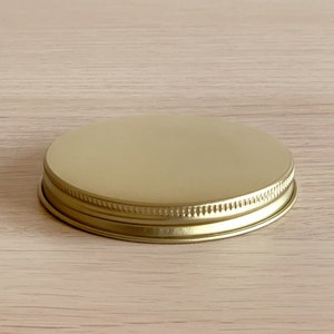May include: A gold-colored metal jar lid with a smooth, flat top and a ridged edge. The lid is circular and sits on a light-colored wooden surface. The lid is designed to seal a jar.