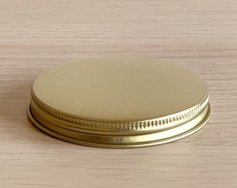 May include: A gold-colored metal jar lid with a smooth, flat top and a ridged edge. The lid is circular and sits on a light-colored wooden surface. The lid is designed to seal a jar.