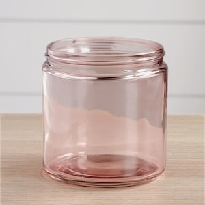 May include: A clear, cylindrical glass jar with a soft pink hue. The jar has a wide opening and a threaded rim. The jar is sitting on a light-colored wooden surface. The background is a white wall.