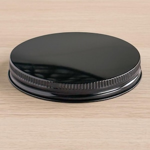 May include: A black, round metal lid with a shiny finish. The lid has a ribbed edge and is designed to fit a jar or container. The lid is shown on a light brown wooden surface.