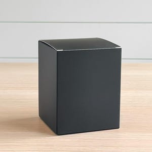 May include: A matte black square box with a silver trim on the top edge. The box is sitting on a light-colored wooden surface. The background is a white wall with horizontal paneling.