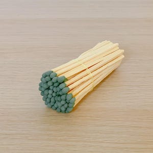 May include: A bundle of wooden matches with green tips, held together by a gold rubber band. The matches are arranged in a fan shape, resting on a light wooden surface.