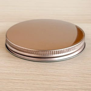 May include: A shiny, metallic jar lid with a silver rim and a smooth, copper-colored top. The lid is round and appears to be made of metal. The rim has a textured, ridged design. The lid is designed to fit on a jar.