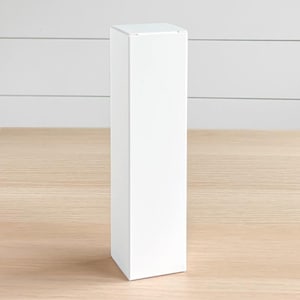 May include: A tall, rectangular white box with a square top and a clean, minimalist design. The box is standing upright on a light-colored wooden surface, with a white wall in the background.