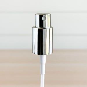 May include: A close-up of a shiny, silver metal perfume or cologne bottle sprayer. The sprayer has a cylindrical body and a small, round nozzle. A white plastic tube extends from the bottom.