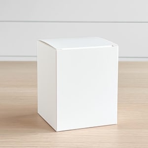May include: A white, square box with a closed lid sits on a light-colored wooden surface. The box is plain and unadorned, with clean lines and a simple design. The background is a white wall with horizontal paneling.