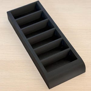 May include: Black plastic organizer with six rectangular compartments. The organizer has a curved edge and is designed to hold small items.