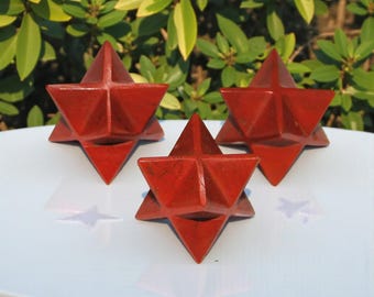 May include: Three red, star-shaped decorative objects with geometric facets. The objects are arranged on a white surface, with a backdrop of green foliage. The stars have a textured appearance and cast subtle shadows.