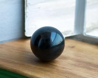 May include: A smooth, black obsidian sphere, polished to a high shine. The sphere is resting on a wooden surface.
