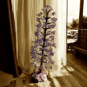 May include: A purple amethyst crystal tree with a wire frame and a natural amethyst base. The tree is standing on a wooden floor in front of a window with white curtains.