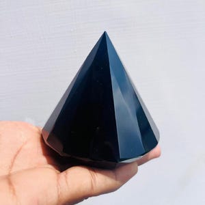 Könnte beinhalten: Ein schwarzer Obsidian-Pyramidenkristall mit einer glatten, polierten Oberfläche. Der Kristall wird in einer Hand gehalten, wodurch seine Größe und Form hervorgehoben werden.