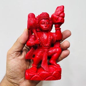 以下が含まれることがあります： 鮮やかな赤色の神像。おそらくハヌマーンで、メイスと山を持っています。この像は、王冠と装飾的な衣服で、複雑に詳細に作られています。像は小さな台座の上にあり、全体的なデザインは象徴的です。