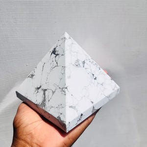 Könnte beinhalten: Eine weiße Pyramide mit grauer Maserung wird in einer Hand gehalten. Die Pyramide hat eine quadratische Basis und vier dreieckige Seiten, die sich in einem Punkt treffen. Der Stein hat ein marmorähnliches Aussehen.