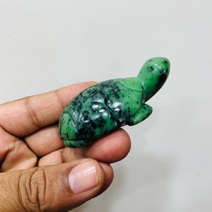 May include: A small, polished green stone carving in the shape of a turtle. The figurine features a vibrant emerald green base color with dark black speckled inclusions throughout the surface, highlighting the shell pattern and the turtle's head.
