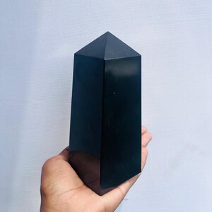 May include: A black obsidian crystal point, also known as a black glass crystal, is a polished, faceted, and pointed crystal. The crystal is approximately 6 inches tall and is held in a person's hand.