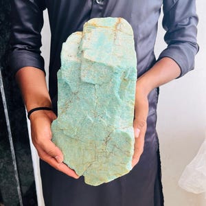 Op de afbeelding: Een groot, ruw stuk amazonietmineraal met een bleke mintgroene kleur, zichtbare kristallijne structuren en natuurlijke bruine aders over het hele oppervlak. De steen heeft een onregelmatige, blokvormige structuur met ruwe, getextureerde randen.