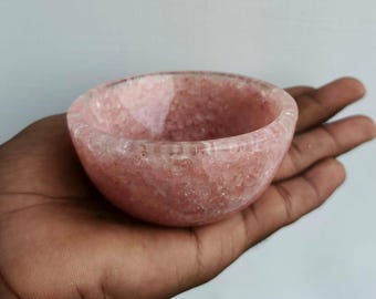 May include: A small, round bowl made of pink stone. The bowl is held in a hand, showcasing its smooth, polished surface and natural color variations. The bowl's interior is visible, revealing its depth and shape.