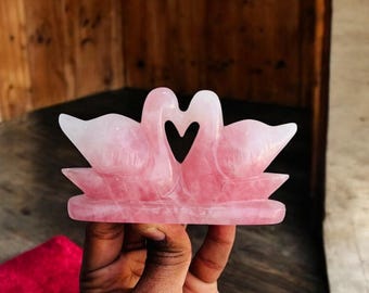 Op de afbeelding: Een roze rozenkwarts sculptuur van twee zwanen met hun nekken in elkaar gevlochten, die een hart vormen.