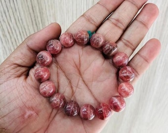 May include: A beaded bracelet featuring round, reddish-pink gemstones. The beads have a translucent quality with darker red and brown inclusions. The bracelet is strung on a green elastic cord and is displayed on a person's open hand.