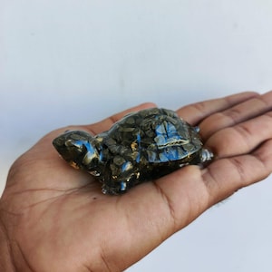 May include: A small, detailed turtle figurine made of a dark, textured material with flecks of gold and blue. The turtle is resting in a person's open hand against a white background. The figurine has a polished, reflective surface.