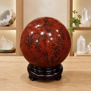 May include: A polished, spherical stone with a deep red base color and scattered black speckles throughout its surface. The sphere is displayed resting on a dark brown, circular wooden stand, showcasing a smooth, reflective finish.
