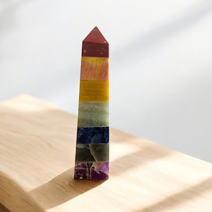 May include: A colorful, layered crystal obelisk. The obelisk features bands of red, yellow, green, blue, and purple. The crystal is standing on a wooden surface.