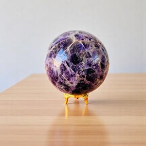Könnte beinhalten: Eine violette Amethystkugel auf einem goldenen Metallständer. Die Kugel ist poliert und hat verschiedene Violetttöne.