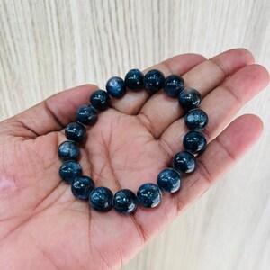 May include: A dark blue beaded bracelet. The bracelet is made of round, polished beads with a reflective surface. The beads are strung together to form a circular shape. The bracelet is held in a hand.