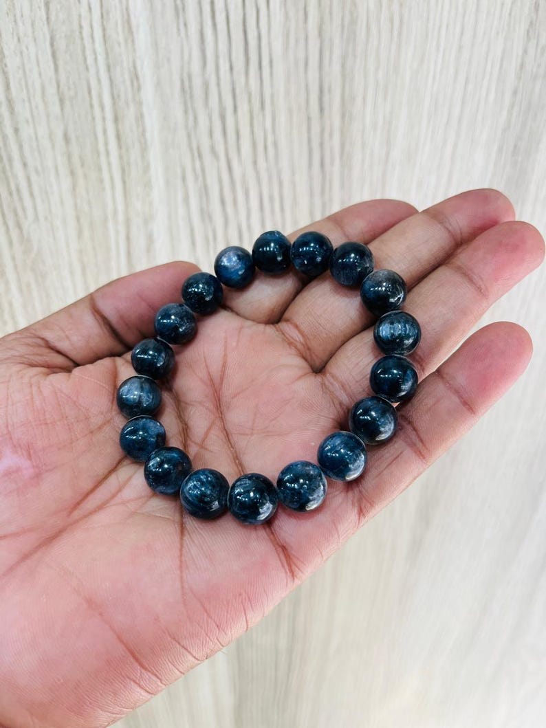 May include: A dark blue beaded bracelet. The bracelet is made of round, polished beads with a reflective surface. The beads are strung together to form a circular shape. The bracelet is held in a hand.