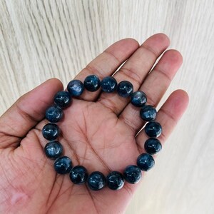 May include: A dark blue beaded bracelet. The bracelet is made of round, polished beads. The beads have a marbled appearance with lighter blue and white inclusions. The bracelet is displayed on a hand.