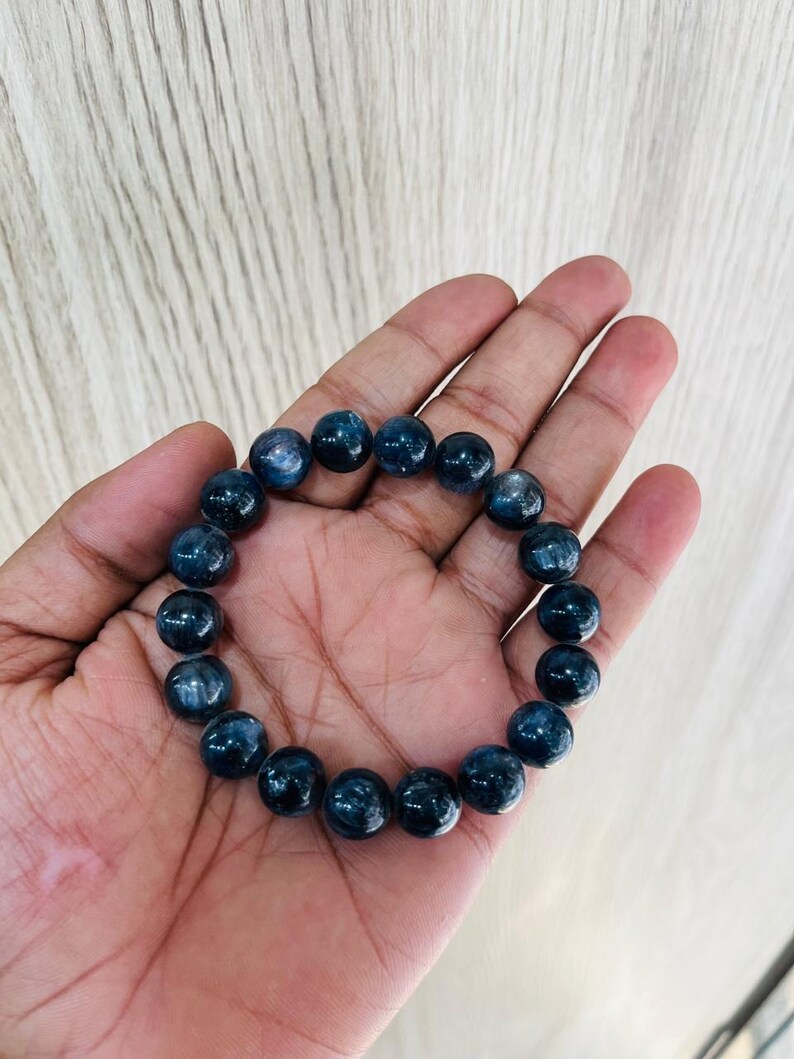 May include: A dark blue beaded bracelet. The bracelet is made of round, polished beads. The beads have a marbled appearance with lighter blue and white inclusions. The bracelet is displayed on a hand.