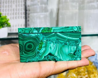 May include: A rectangular, hinged box made of polished malachite, a vibrant green mineral with swirling patterns. The box is held in a hand, showcasing the intricate natural designs and the rich colour variations of the stone. The box is approximately 10 cm long.