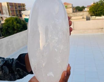 May include: A large, clear quartz crystal egg is held in a person's hands. The egg-shaped crystal is translucent with visible internal patterns. The background shows a rooftop and buildings under a clear sky.