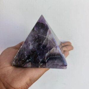 Könnte beinhalten: Eine pyramidenförmige Amethystkristall. Der Kristall hat eine tiefviolette Farbe mit helleren violetten und weißen Adern. Die Pyramide wird in einer Hand vor einem weißen Hintergrund gehalten. Die Pyramide ist etwa 10 cm hoch.