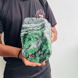 May include: A large, rough-cut mineral specimen featuring a vibrant mix of bright green, deep black, and small magenta-colored crystalline inclusions. The stone has a textured, uneven surface with distinct color banding and a raw, natural appearance.