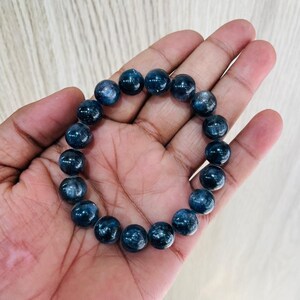May include: A dark blue beaded bracelet. The bracelet is made of round, polished beads. The beads have a speckled appearance and are strung together on an elastic band. The bracelet is shown on a person's hand.