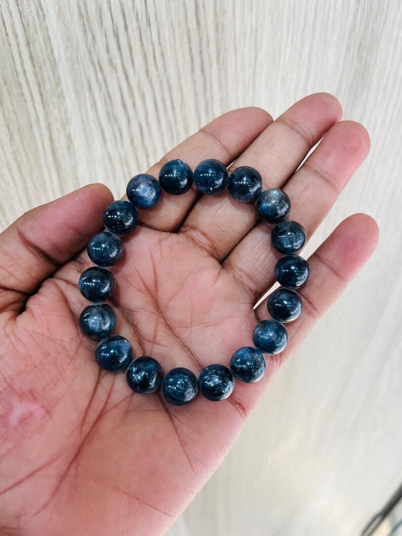 May include: A dark blue beaded bracelet. The bracelet is made of round, polished beads. The beads have a speckled appearance and are strung together on an elastic band. The bracelet is shown on a person's hand.