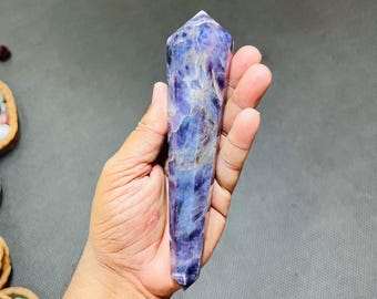 May include: A polished, elongated crystal point with a faceted top. The crystal features a blend of purple and blue hues, with streaks of lighter shades throughout. The crystal is held in a hand against a gray background.