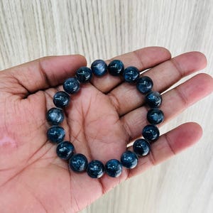 May include: A dark blue beaded bracelet. The bracelet is made of round, polished beads. The beads have a speckled appearance and are strung together on an elastic band. The bracelet is shown resting on a hand.