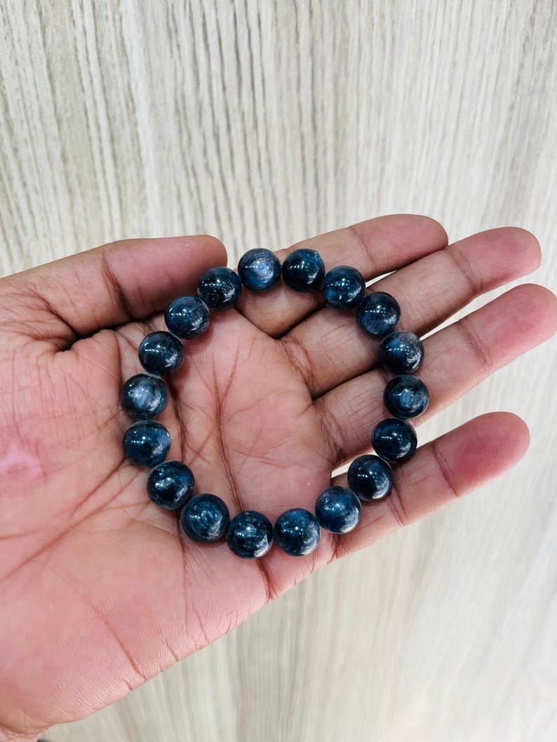 May include: A dark blue beaded bracelet. The bracelet is made of round, polished beads. The beads have a speckled appearance and are strung together on an elastic band. The bracelet is shown resting on a hand.