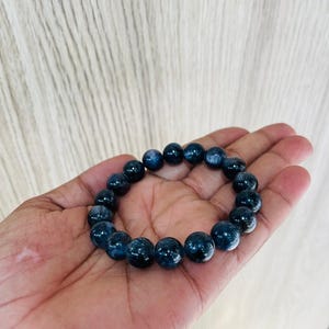 May include: A dark blue beaded bracelet. The bracelet is made of round, polished beads. The beads have a marbled appearance with lighter blue and white streaks. The bracelet is held in a hand.