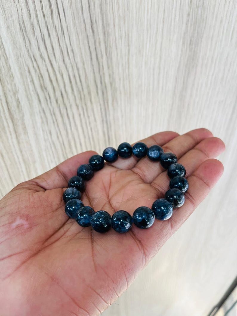 May include: A dark blue beaded bracelet. The bracelet is made of round, polished beads. The beads have a marbled appearance with lighter blue and white streaks. The bracelet is held in a hand.