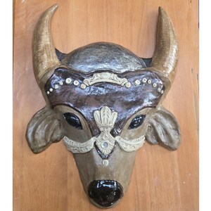May include: A brown and gray ceramic bull mask with ornate details, including a silver-colored nose ring and decorative accents around the eyes.